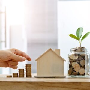 Hand symbolically places coins next to a model of a house. a jar of coins is next to it with leaves growing out of it. 